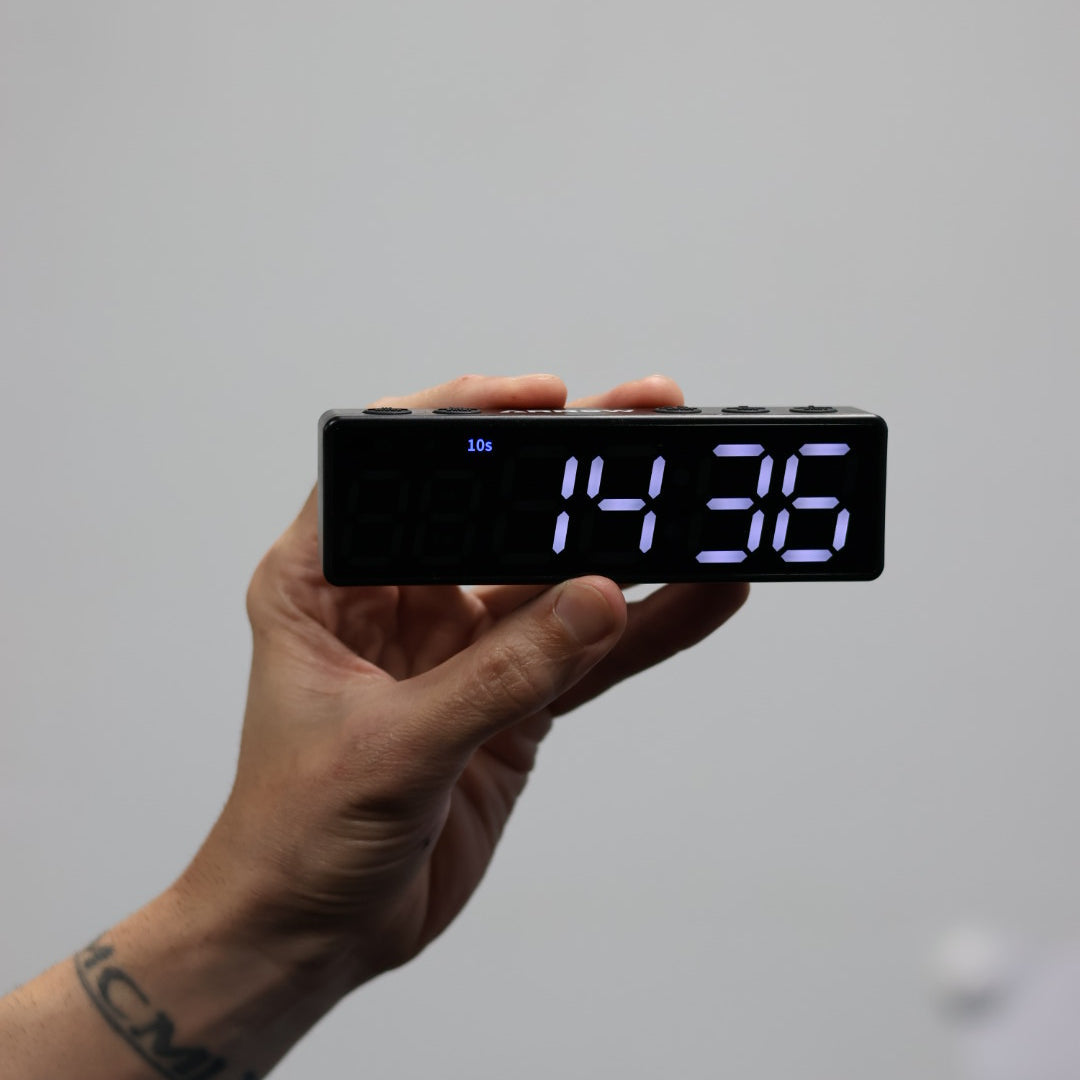Hand holding a digital clock displaying the time '14:35' on a plain background