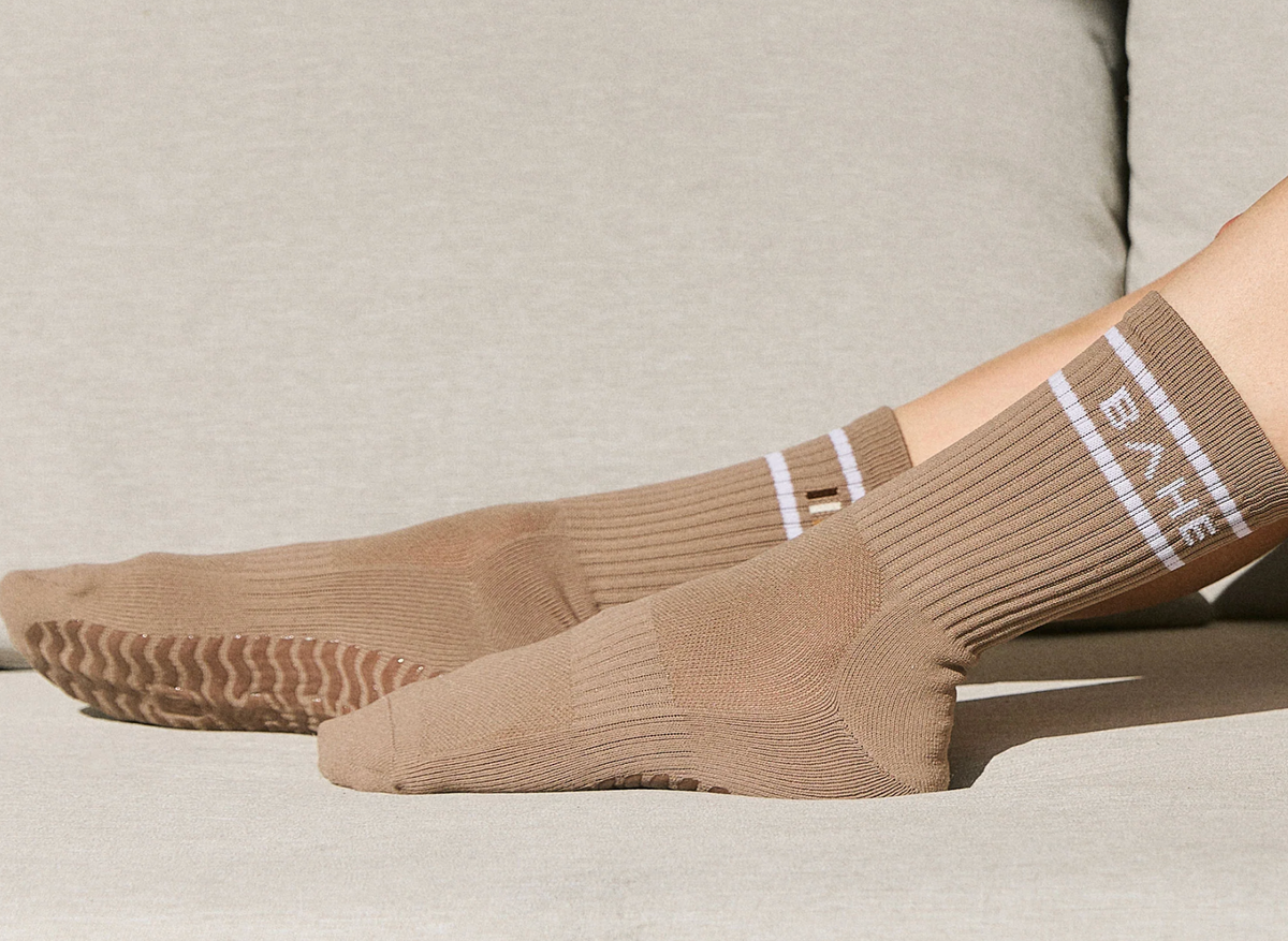 A pair of taupe-brown ribbed crew socks with white stripe details and the brand name 'BAE' printed vertically, shown on a person's feet resting on a light beige sofa.